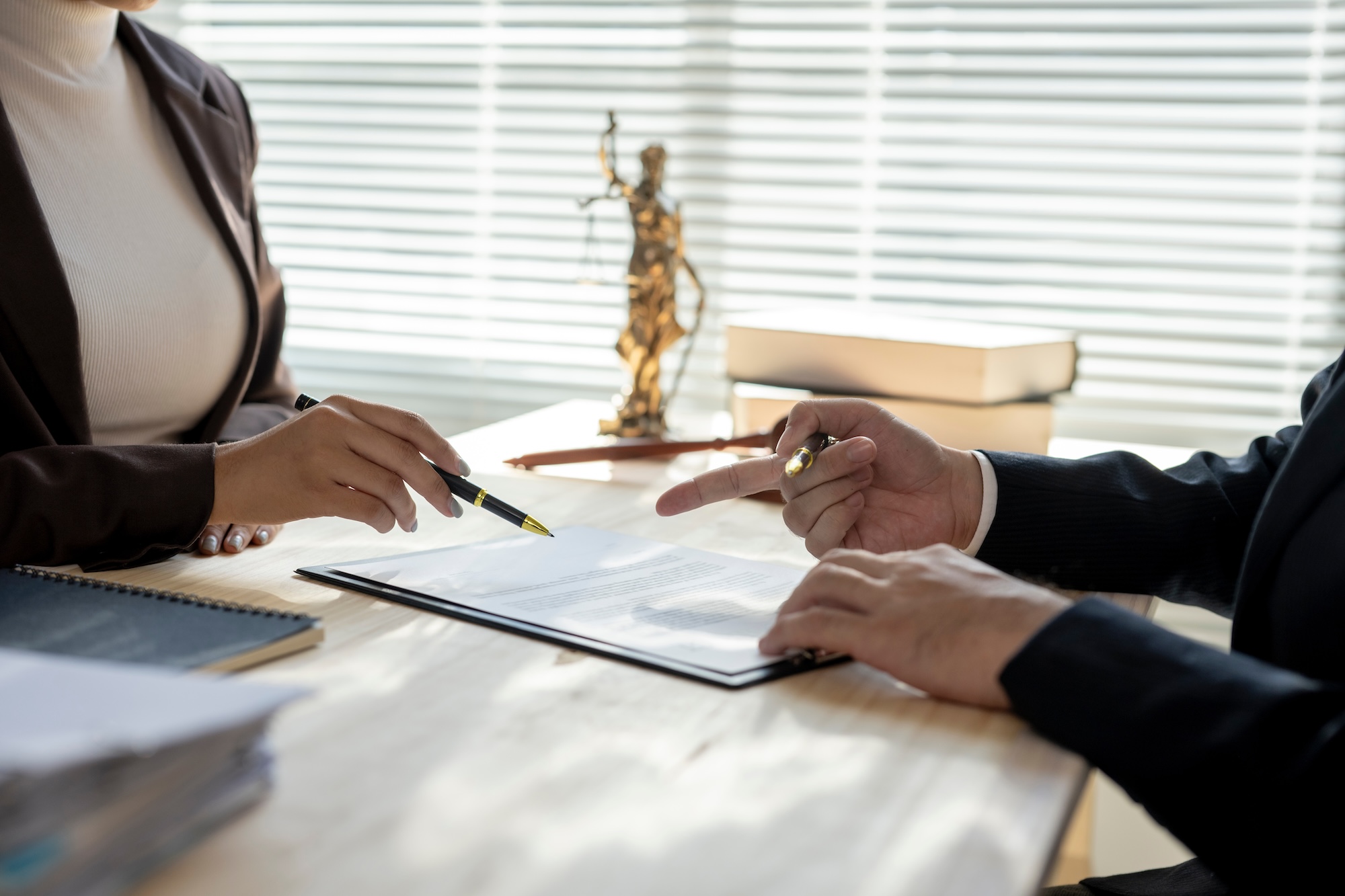 Paperwork on a desk with two people on either side of the desk with one person holding a pen directing the other to sign
